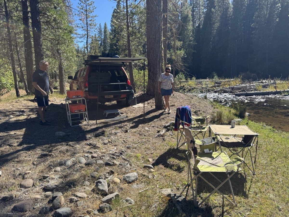Tan SUV with awning and kitchen setup by forest stream