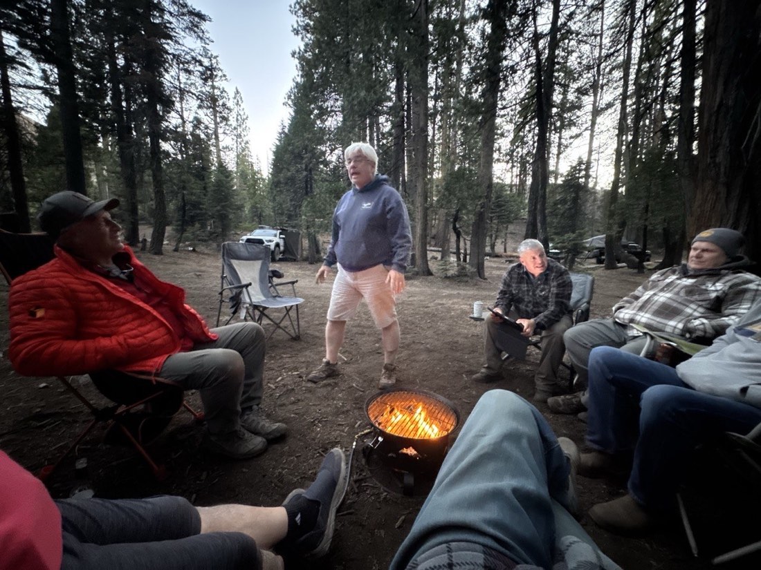 East Bay Overland members seated around a propane fire pit at dusk as the trip leader addresses the camp circle in Tahoe National Forest